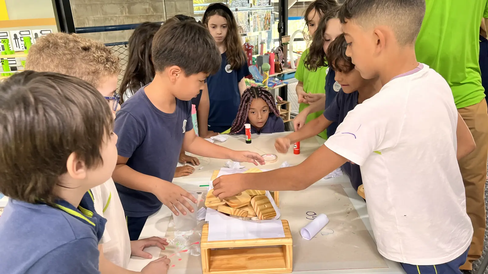 Estudantes reunidos em torno de uma mesa, trabalhando juntos com papel, cola e peças de madeira.