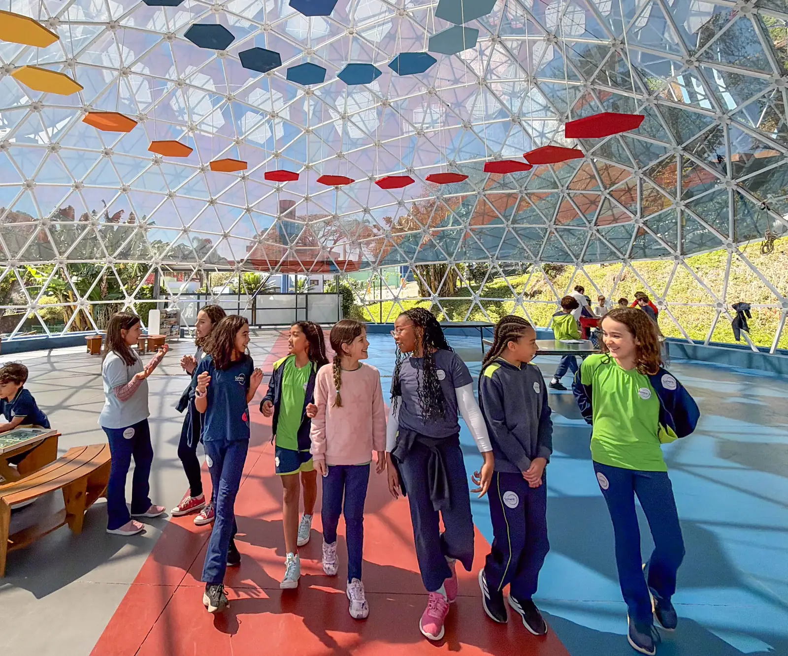 Estudantes de uniforme Sphere conversam e caminham dentro da geodésica de vidro, sob painéis hexagonais coloridos.