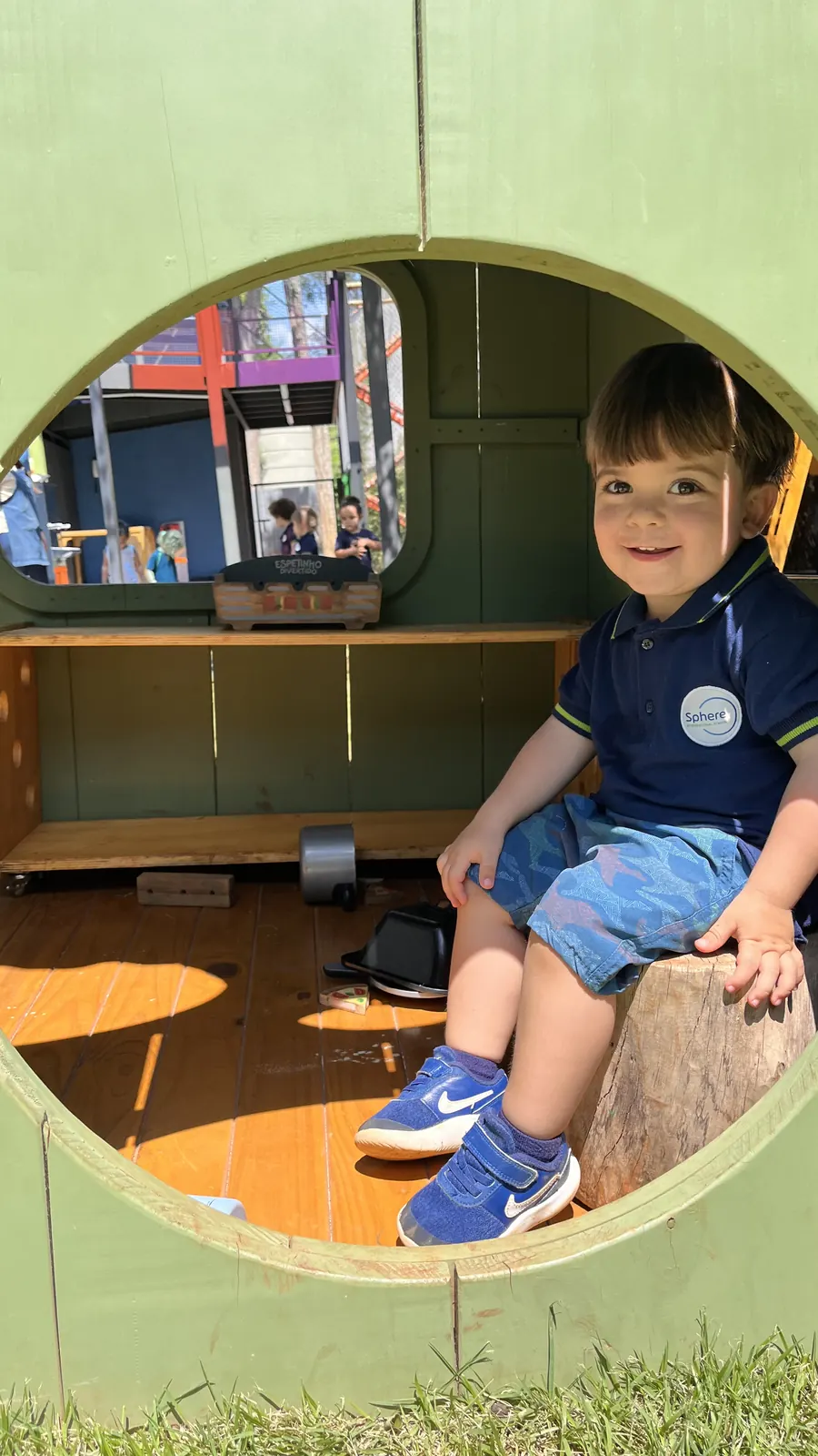 Criança da Educação Infantil, com uniforme Sphere, sorri dentro de uma casinha de madeira no parque.