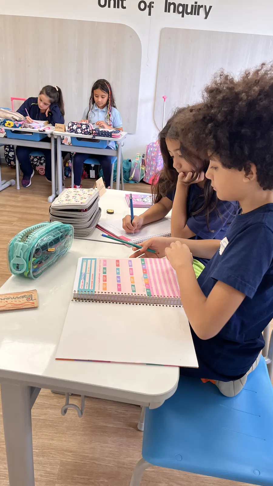 Estudantes dos Anos Iniciais escrevem em seus cadernos, sob a placa Unit of Inquiry.
