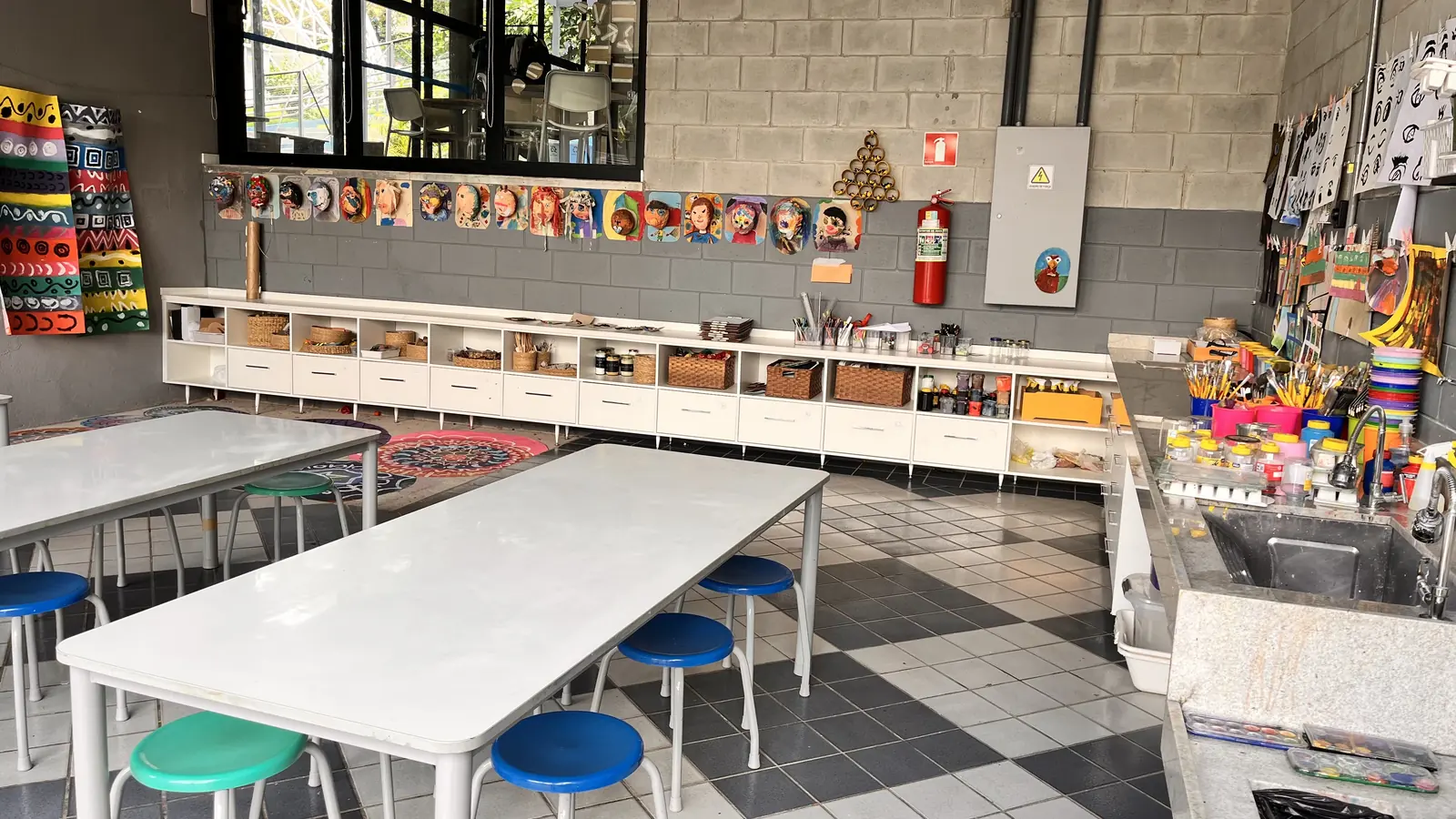 Ateliê de artes com máscaras feitas pelos estudantes na parede, estantes com materiais e mesas com bancos coloridos.