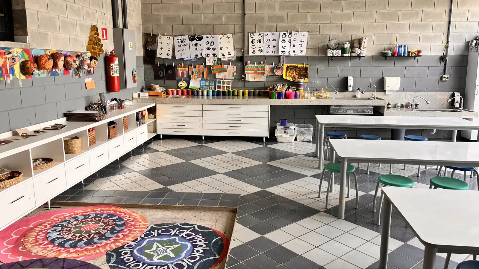 Ateliê de artes com mesas brancas, bancos coloridos, pias, desenhos dos estudantes pendurados e mandalas pintadas no chão.