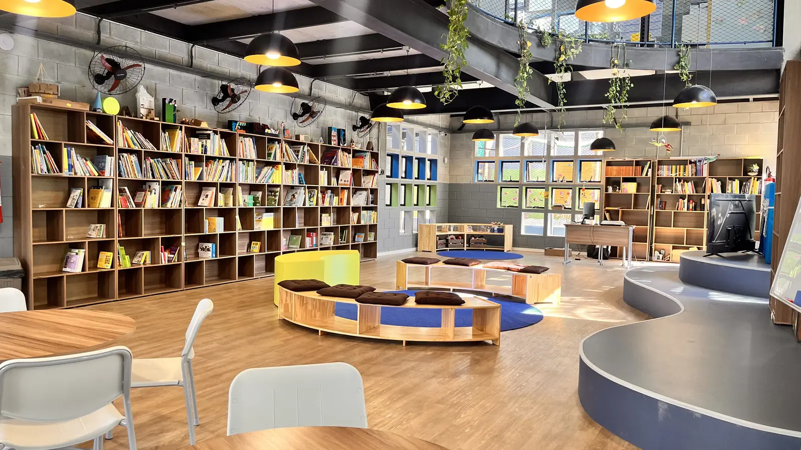 Biblioteca com estantes de madeira cheias de livros, bancos em curva com almofadas e luminárias pendentes.